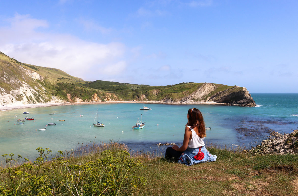 Woman in white top sitting on the cliff above Lulworth Cove watching the fishing boats