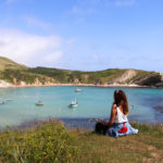 Woman in white top sitting on the cliff above Lulworth Cove watching the fishing boats